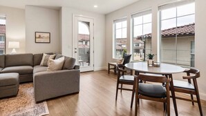 Een moderne woonkamer met een grijze hoekbank, een eettafel met houten stoelen en grote ramen met uitzicht op de omgeving.
