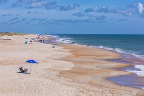 Uma extensa praia de areia com ondas suaves, céu claro e algumas pessoas e guarda-sóis.