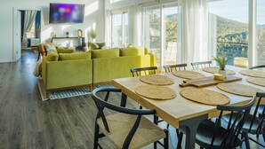 A spacious living area with a sofa, dining table with placemats, and a TV mounted on the wall. - Sunrise+Sunset views - HotTub / Firepit / RecRoom / BBQ / Yoga studio /Treadmill (Central City)