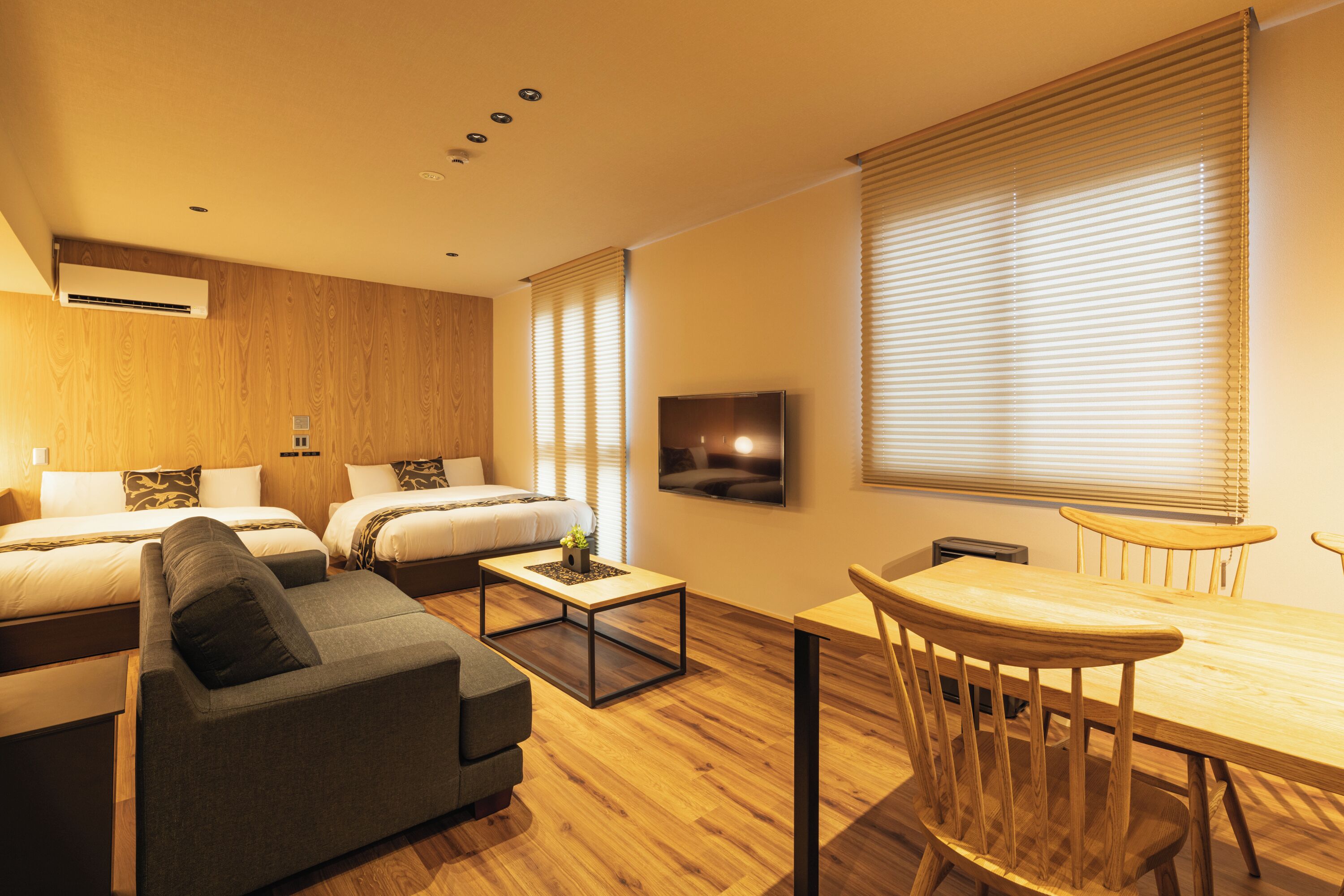 A modern hotel room with two beds, a sofa, a dining table with chairs, and a TV mounted on the wall.