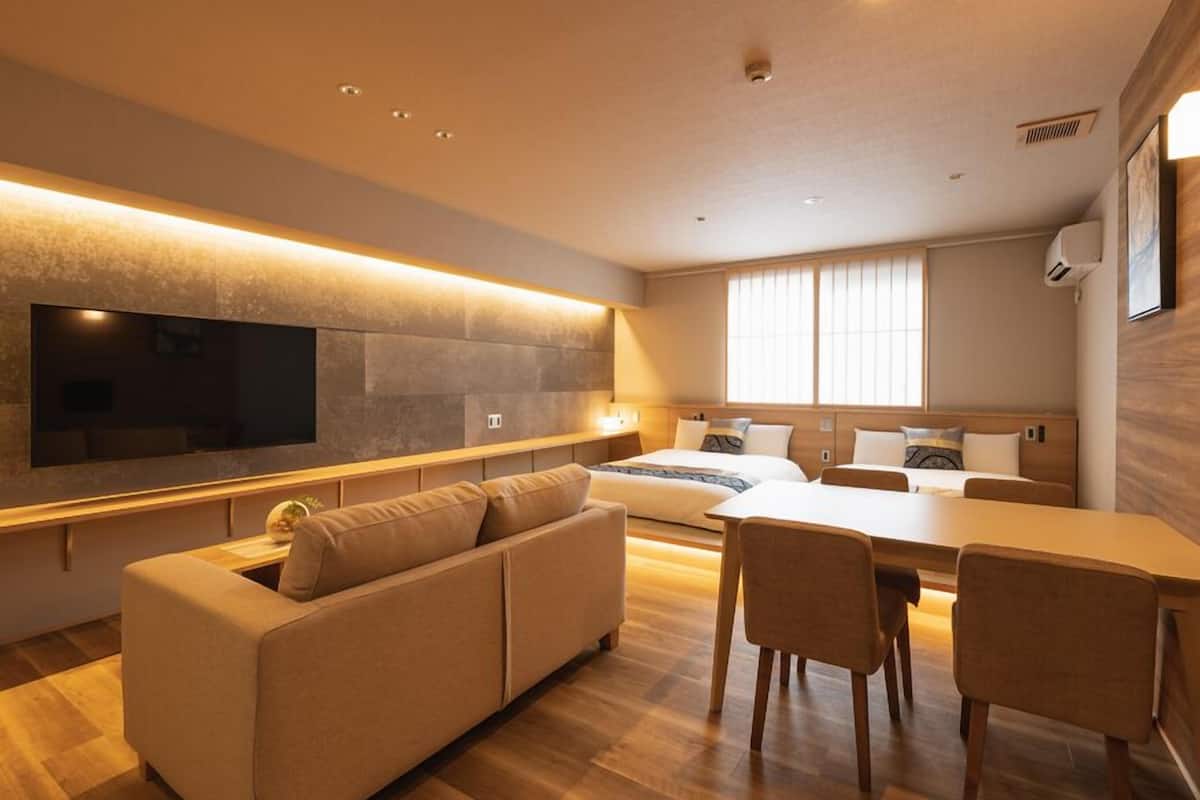 A modern hotel room with a sofa, dining table, chairs, a TV, and a large window.
