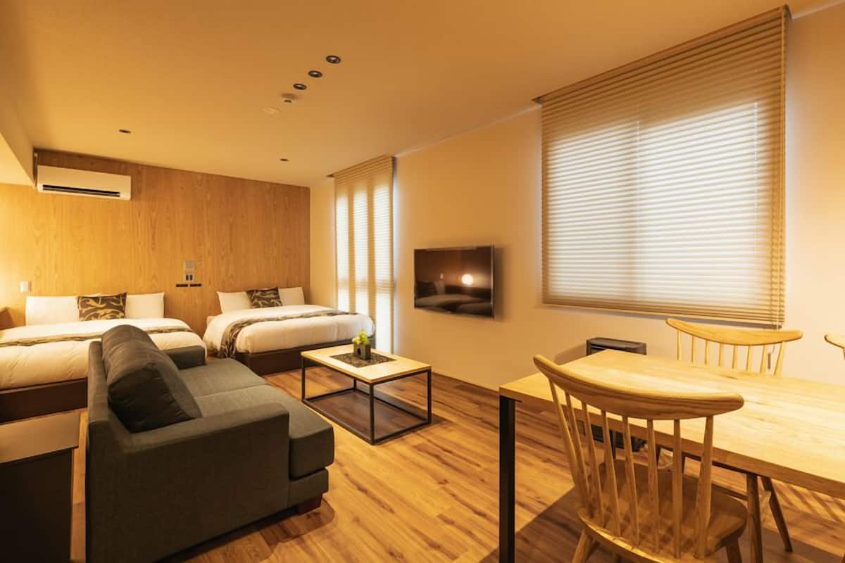 A modern hotel room with a sofa, two beds, a TV, a dining table, and wooden blinds.