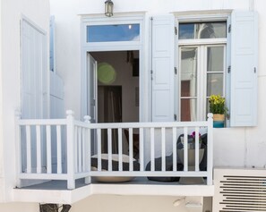 A white balcony with a white railing, a potted plant with yellow flowers, and a window with blue shutters. - Seablue Town Maisonette (Mykonos)
