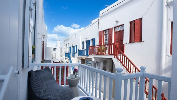 A narrow street with white buildings, red railings, and a balcony with a cushion and a vase.