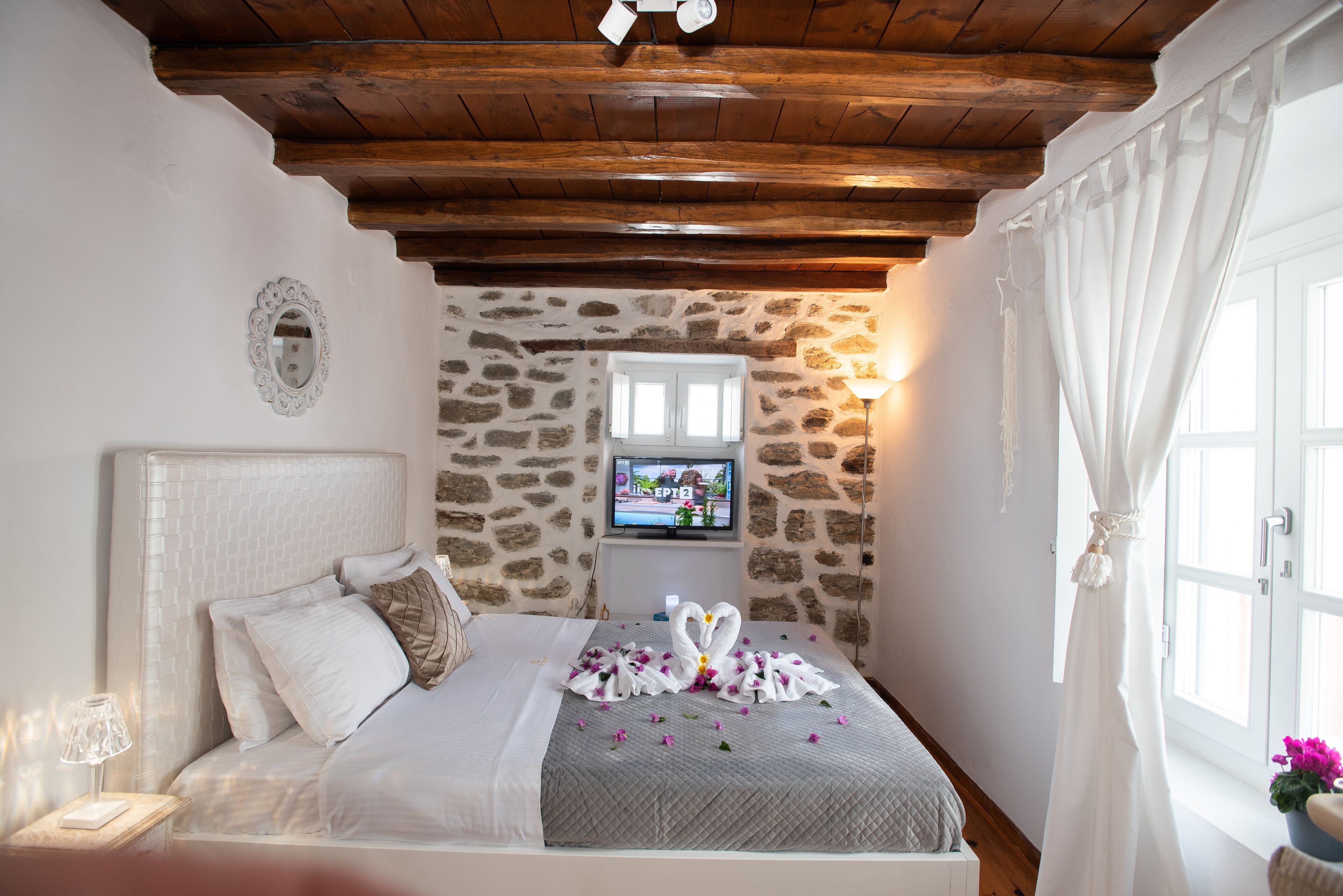 A bedroom with a stone wall, a bed with a white headboard, a TV on a stand, and a window with curtains.