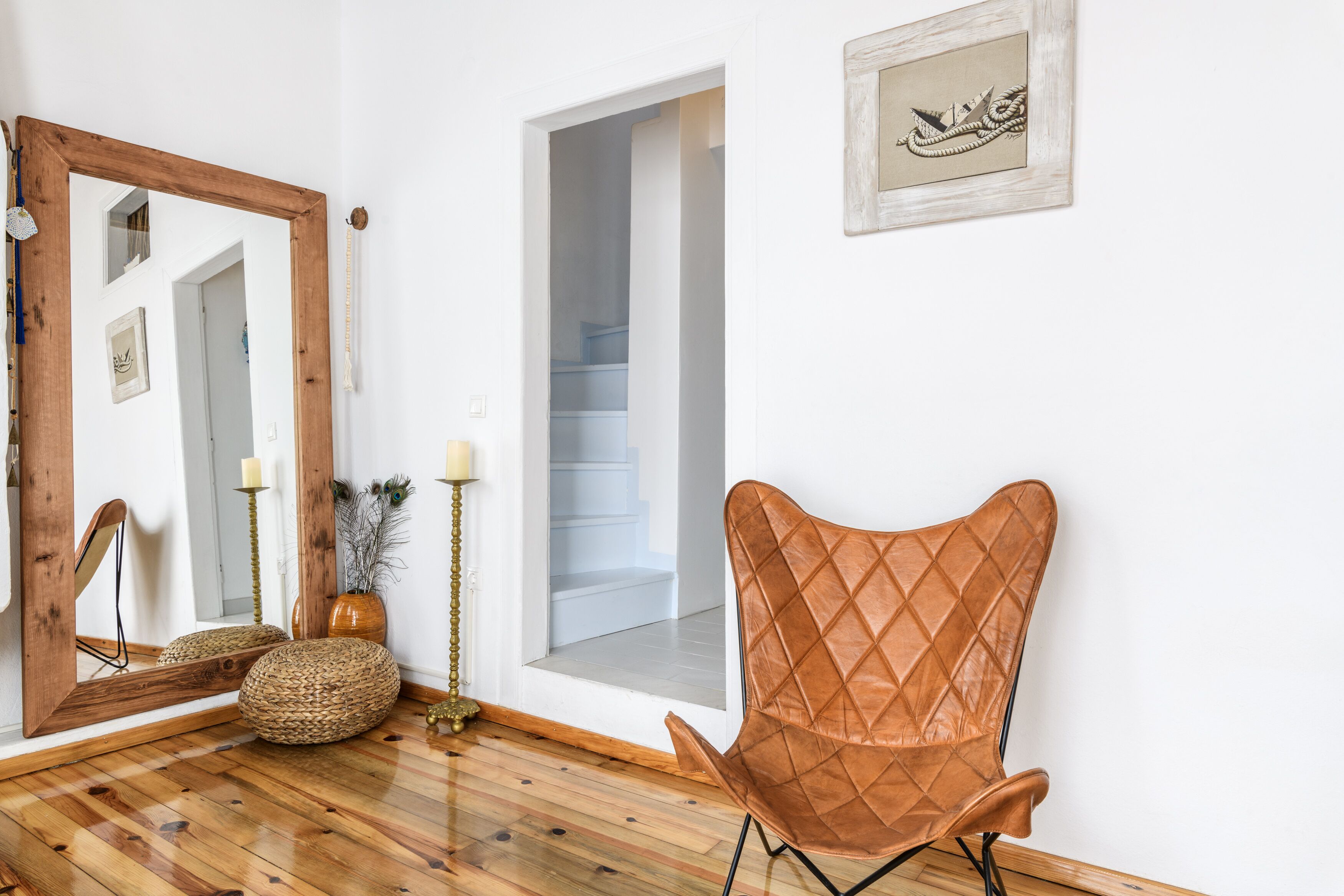 Una habitación con piso de madera, un sillón de cuero marrón, un espejo, un florero con plantas secas y un cuadro en la pared.