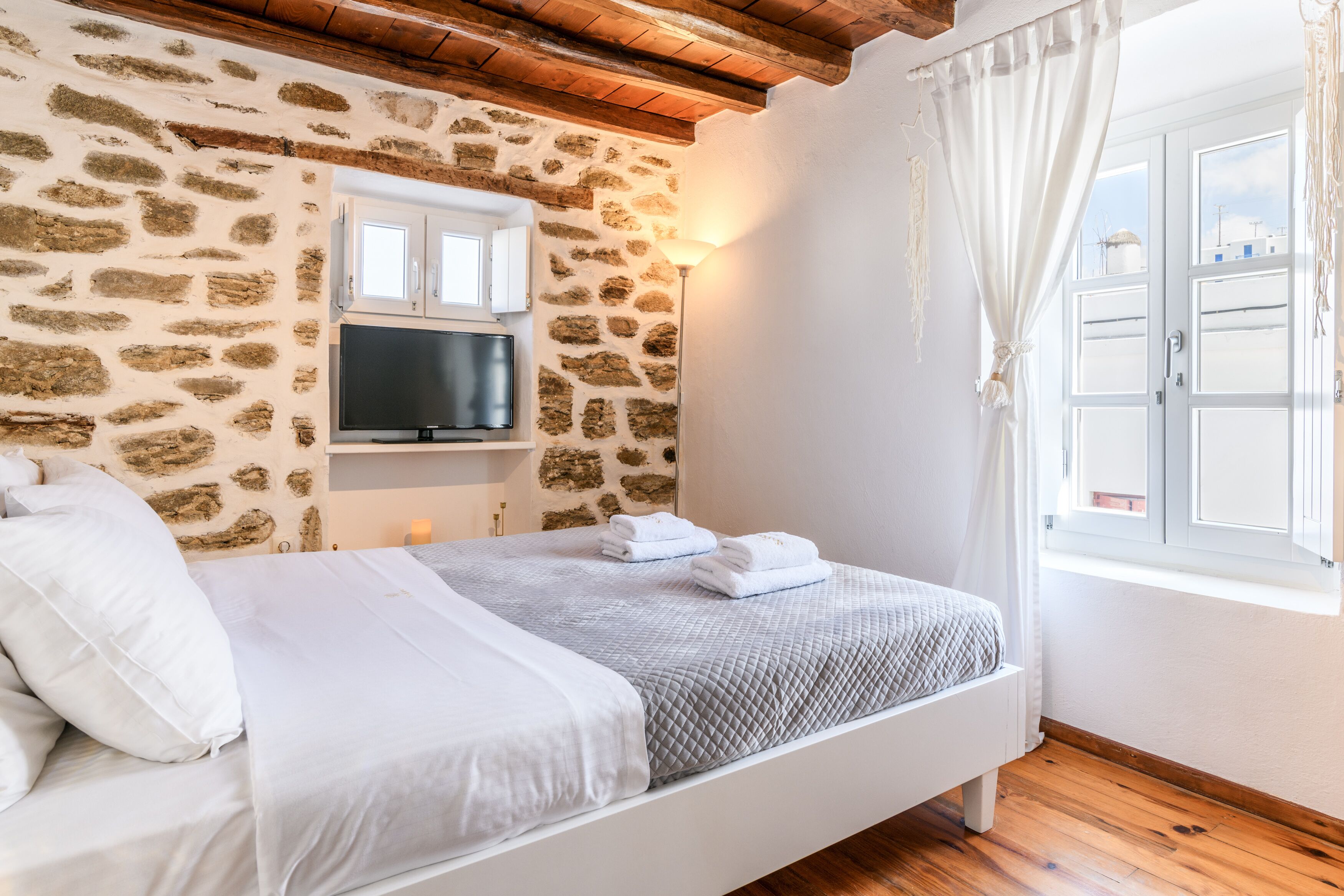 A bedroom with a stone wall, a flat-screen TV, a bed with white bedding, and a window with curtains.