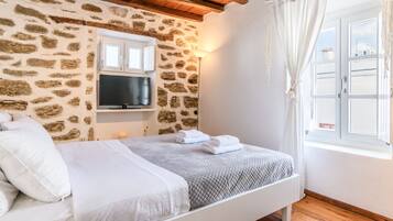 A bedroom with a stone wall, a flat-screen TV, a bed with white bedding, and a window with curtains.