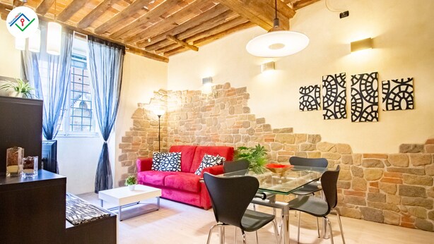 A cozy living area with a red sofa, a glass dining table, and a stone wall. - Antica Piazza Apartment (Lucca)