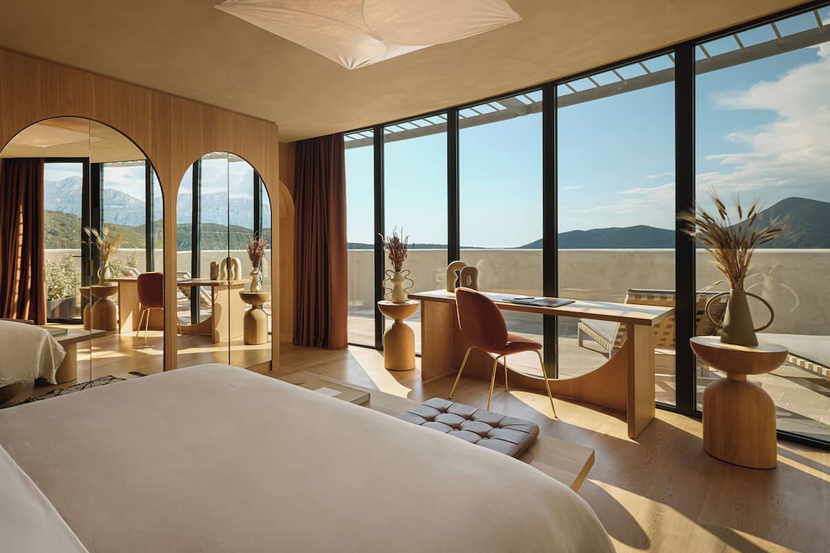 A modern hotel room with a large bed, a desk, and a view of mountains through large windows.