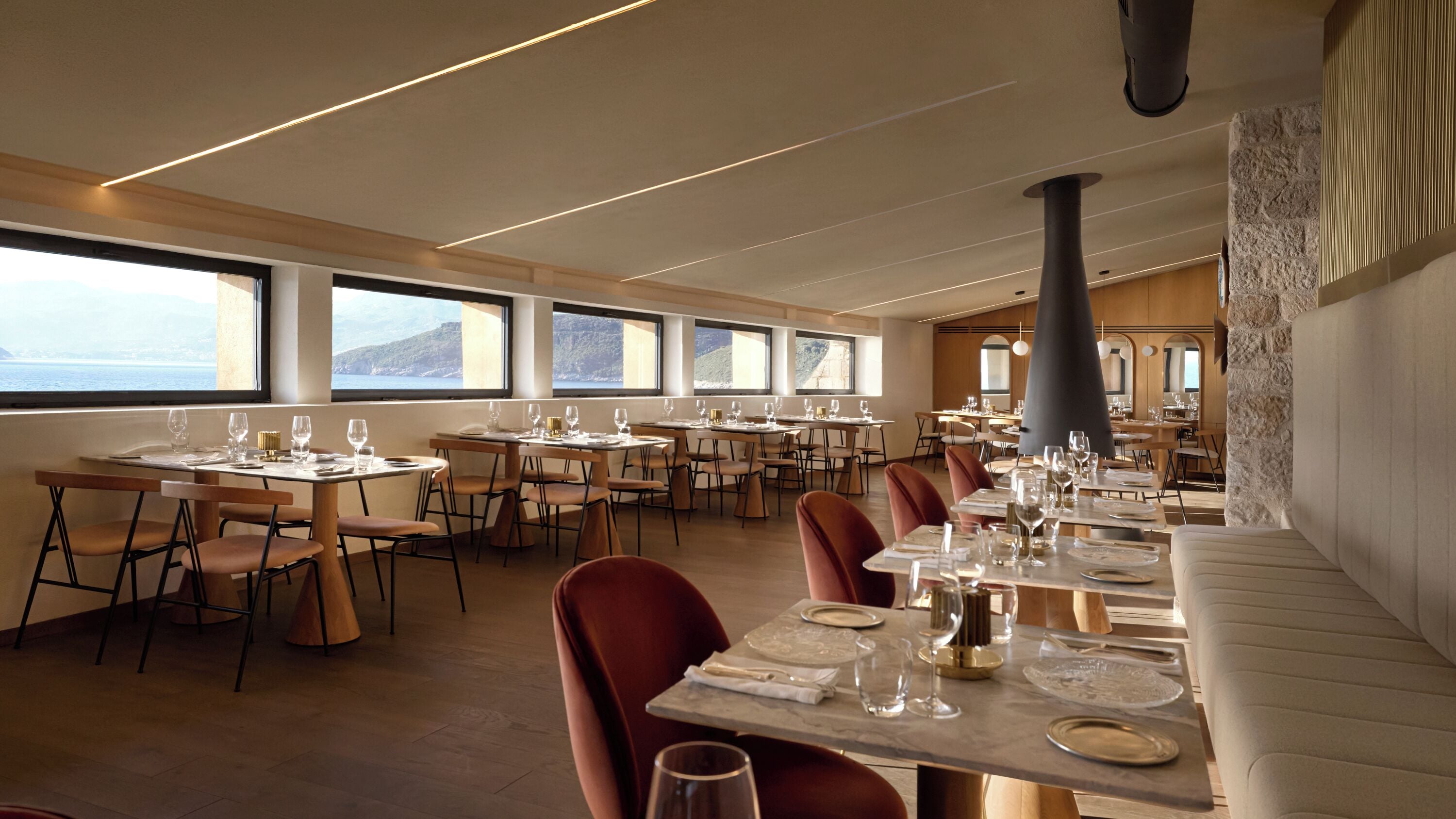 Un restaurante con varias mesas y sillas, grandes ventanales con vista al mar y una pared de piedra en un lateral.
