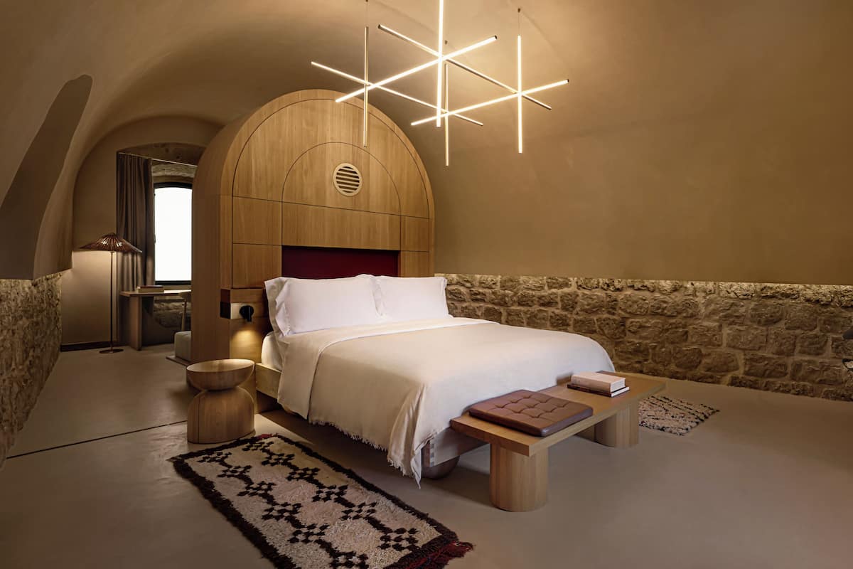 A bedroom with a large bed, a wooden headboard, a stone wall, a modern light fixture, and a small table with books.