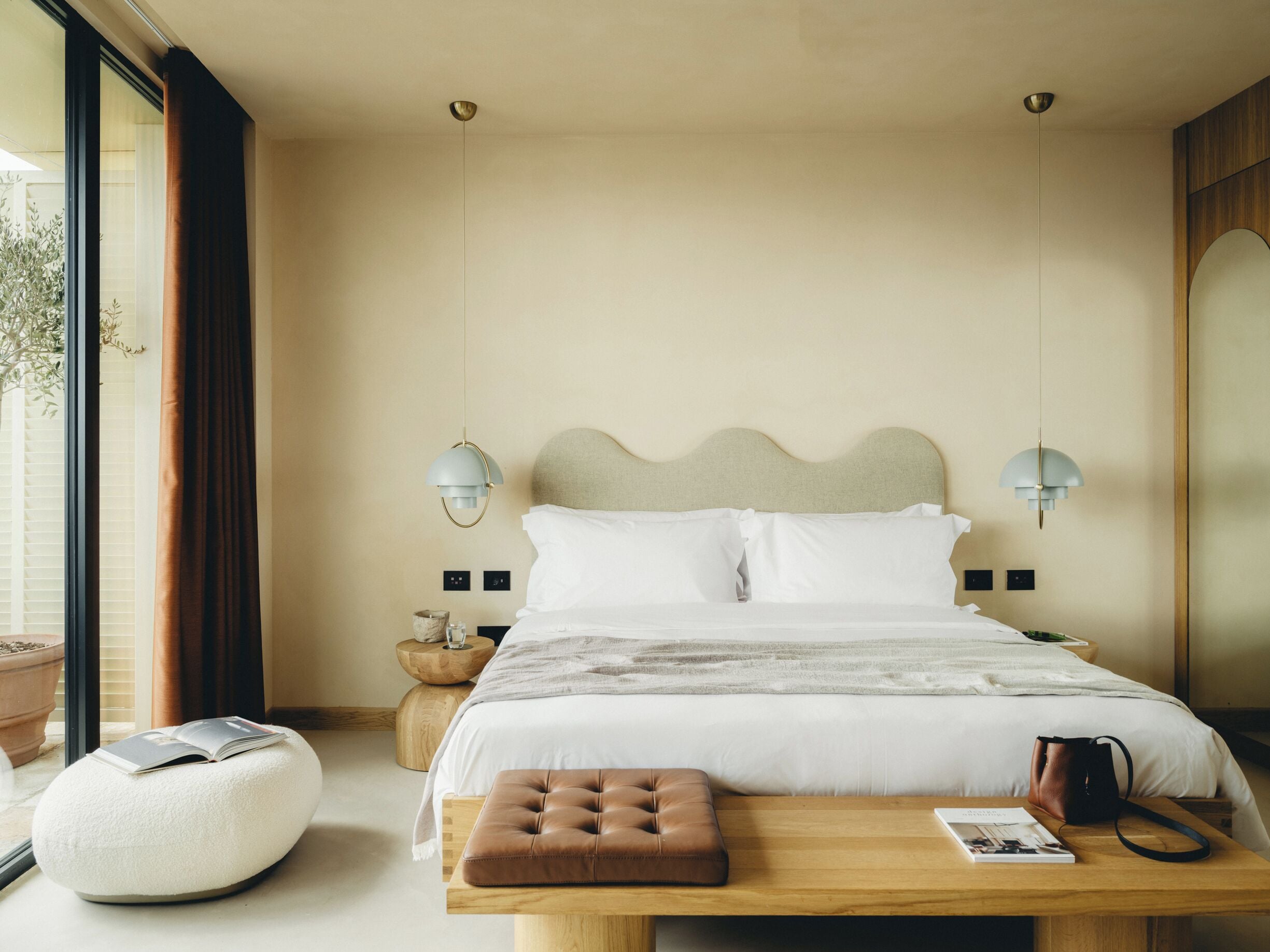 A neatly arranged bedroom with a bed, bedside lamps, a wooden nightstand, a reading bench, and a potted plant.