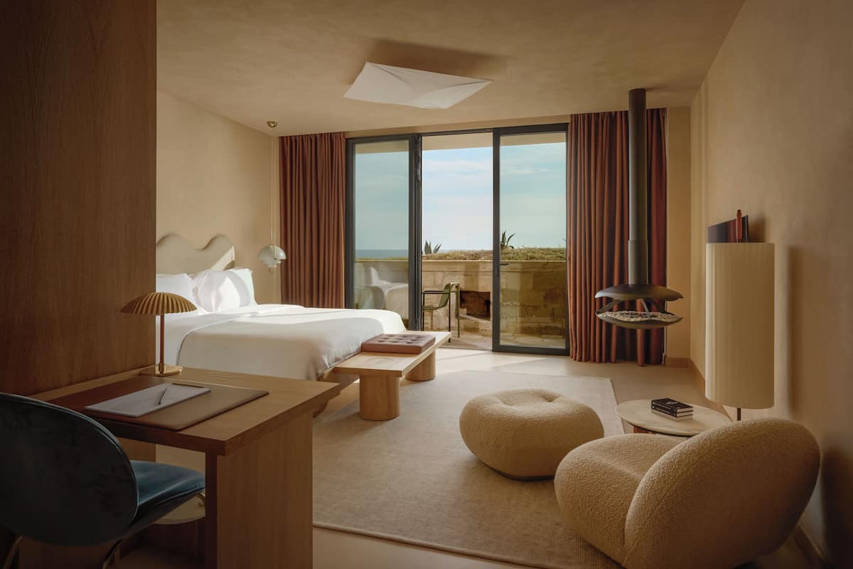 A hotel room with a bed, desk, chair, ottoman, and a fireplace.