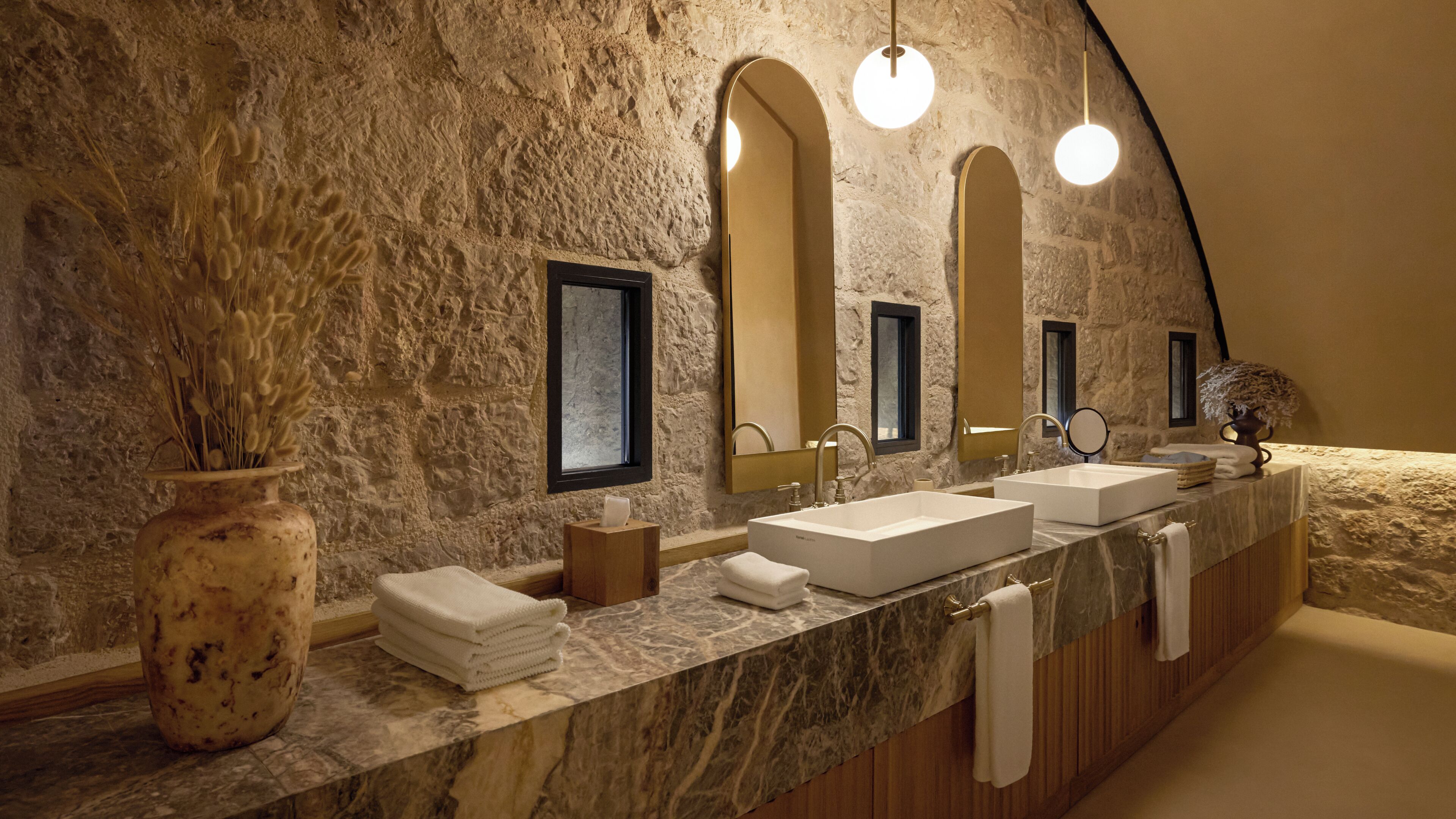 A bathroom with a stone wall, a marble countertop, two sinks, and a large mirror.