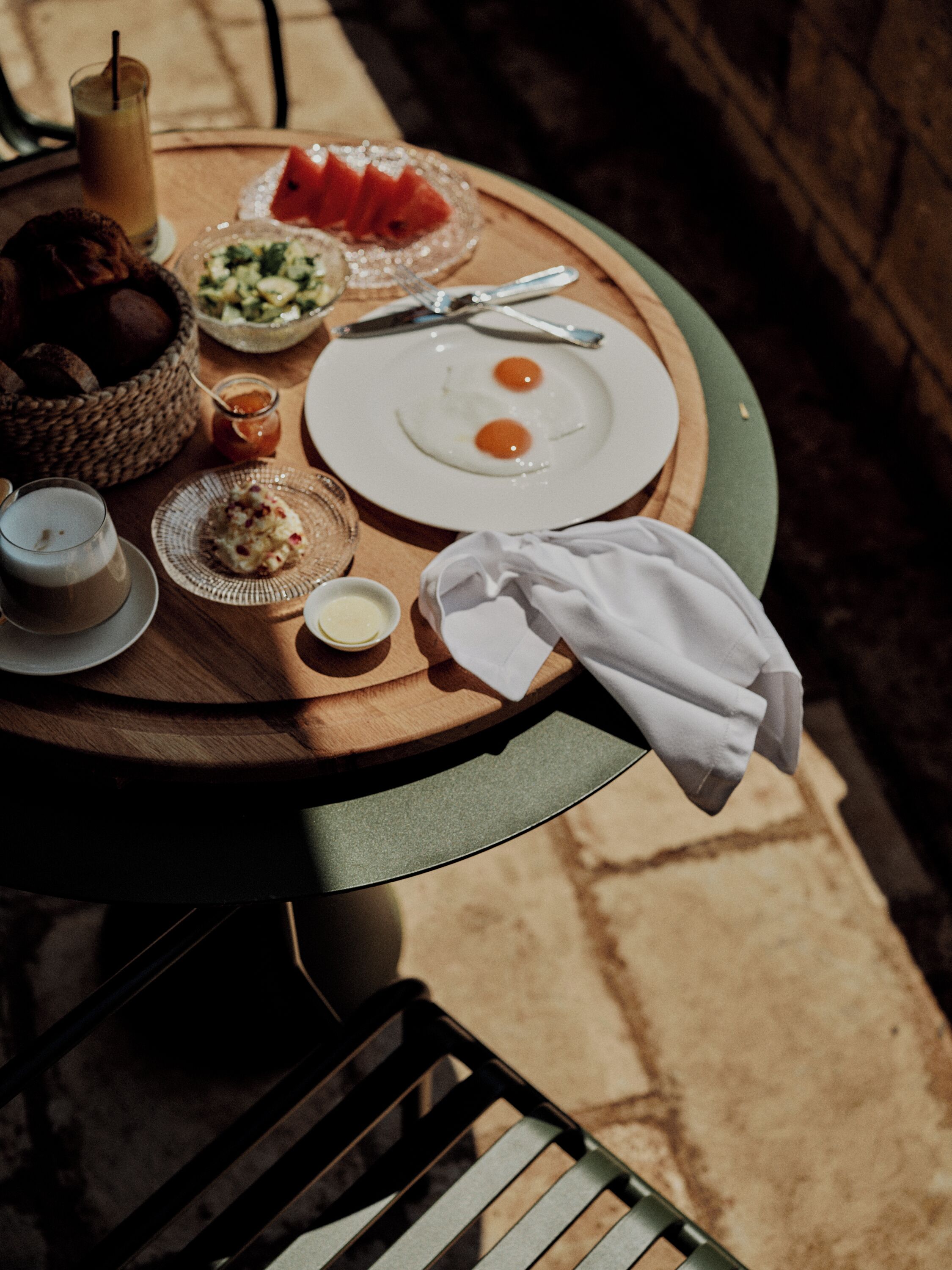 A round wooden table with a white cloth, a basket of bread, a plate with two eggs, a glass of a beverage, a jar of jam, and a napkin.