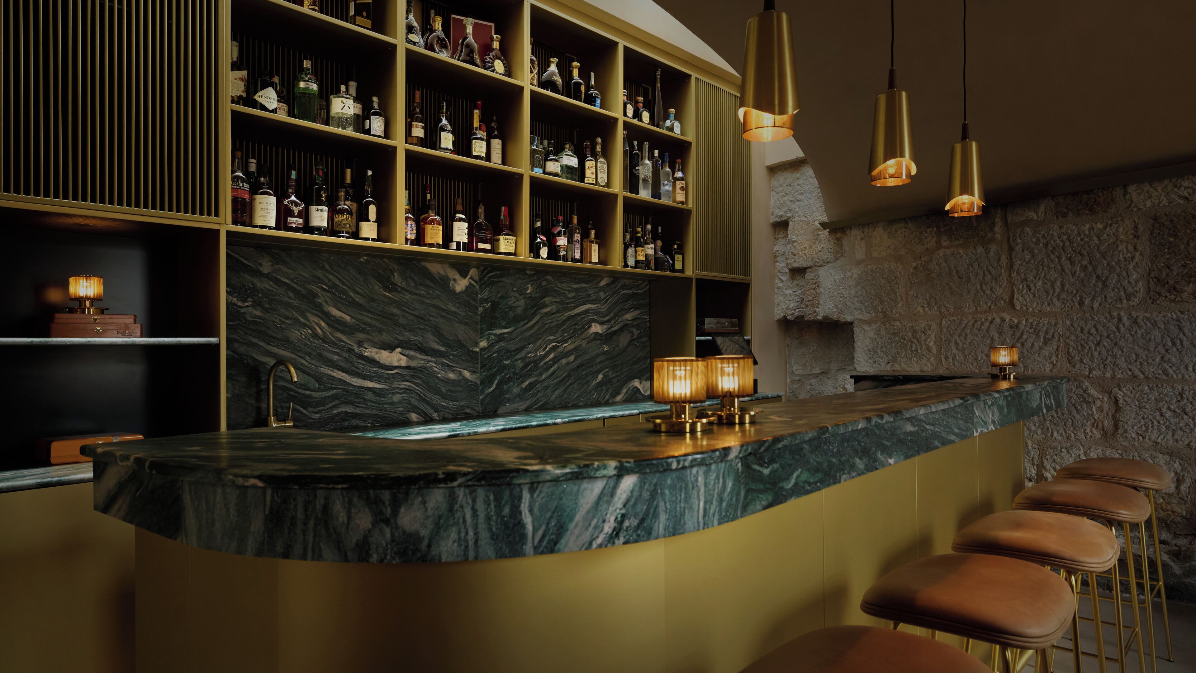 Uma área de bar com bancada de mármore, banquetas, luminárias pendentes e uma parede com garrafas de bebidas.