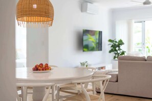 Un coin repas avec une table et des chaises blanches, un luminaire suspendu en osier, une assiette de fruits, un téléviseur accroché au mur et un canapé à l’arrière-plan.