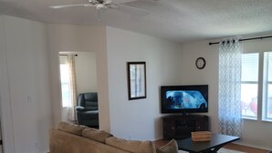 A living room with a beige sofa, a grey armchair, a TV on a stand, and a ceiling fan.