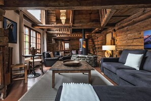 Een rustiek interieur met zichtbare houten balken, een stenen openhaard, lederen meubels en een groot raam dat natuurlijk licht binnenlaat.