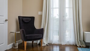 Una habitación con un sillón oscuro, una mesita redonda y una ventana con cortinas traslúcidas.