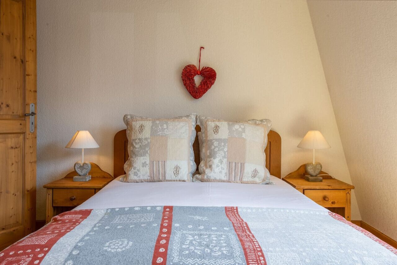 A bedroom with a bed, two nightstands, a heart-shaped decoration on the wall, and a wooden door.