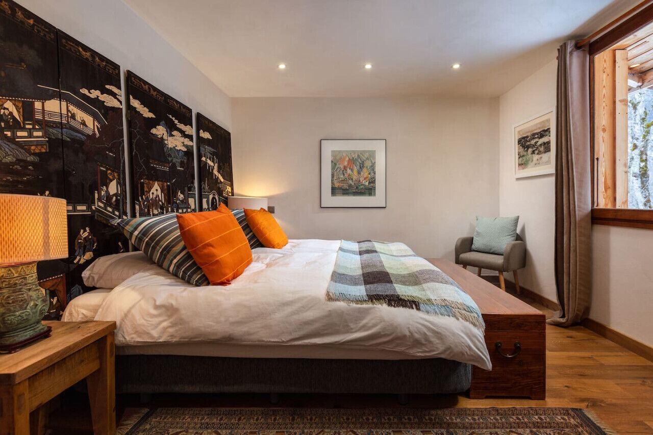 Un dormitorio con una cama, almohadas, una silla, una lámpara, obras de arte en la pared y una ventana.