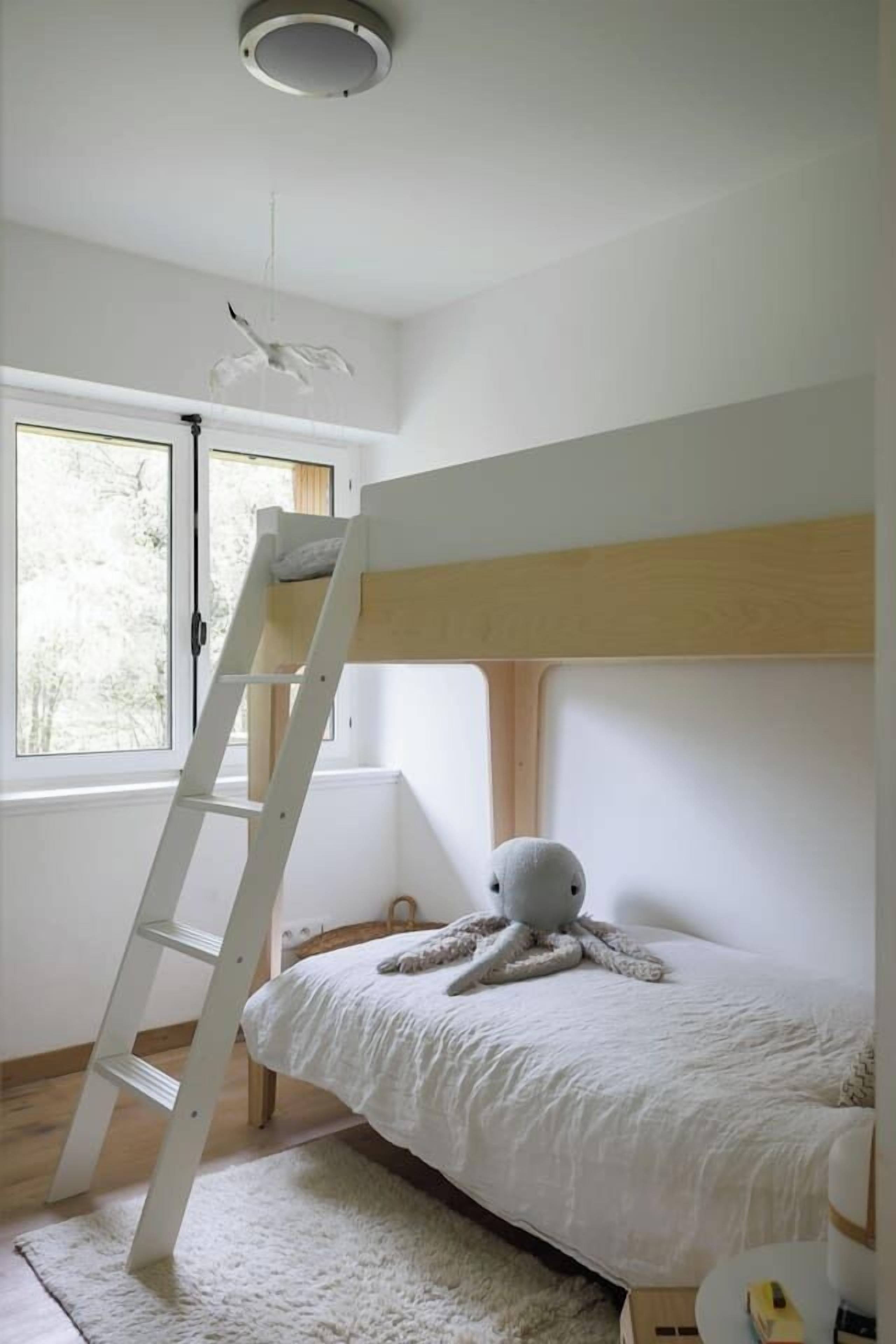 Ein Schlafzimmer mit Etagenbett, einer Stehleiter, einem Kuscheltier, einem weißen Teppich und einem Fenster mit Blick auf Bäume.
