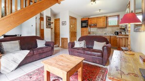 A spacious living area with two leather sofas, a wooden coffee table, and a red lamp. A kitchen with wooden cabinets and a staircase is visible in the background.