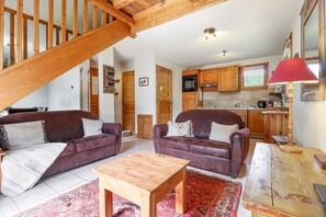 A spacious living area with two leather sofas, a wooden coffee table, and a red lamp. A kitchen with wooden cabinets and a staircase is visible in the background. - Amethyste · 2BR Apart Authentic | Mt Blanc View | Free Parking (Les Houches)