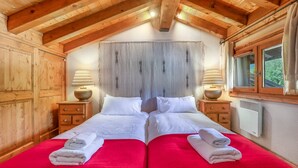 A double room with wooden beams, two beds with red bedding, white pillows, bedside lamps, and a window with curtains.