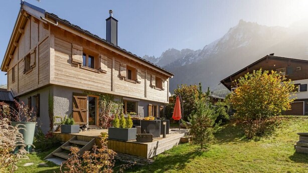 Un chalet en bois avec une cheminée, un petit jardin et une vue sur les montagnes en arrière-plan.