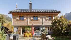 Une maison en bois à deux étages, dotée d’un patio, d’une table avec des chaises et d’un petit jardin.