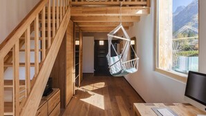 Une pièce de style loft avec un escalier en bois, un fauteuil suspendu, un bureau avec un ordinateur et une grande fenêtre donnant sur la montagne.