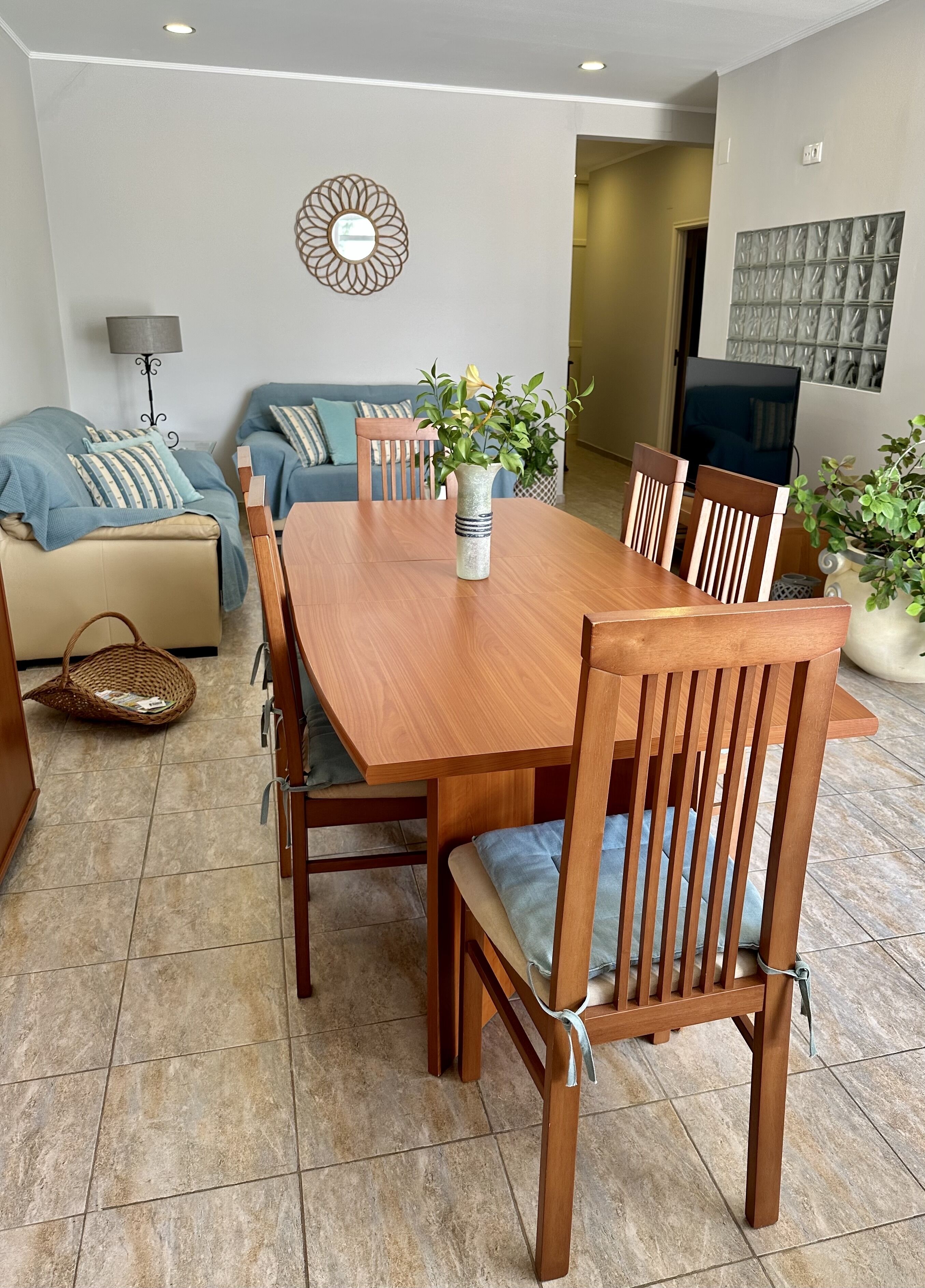 Un coin repas avec une table et des chaises en bois, un vase de fleurs, un canapé et un téléviseur en arrière-plan.