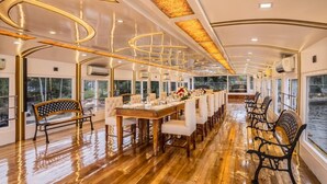 A dining area with a long wooden table, white upholstered chairs, and a bench. - Premium Houseboat (Ambalapuzha)
