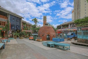 Una plaza al aire libre con un horno de ladrillo, zonas de estar y varios edificios al fondo.