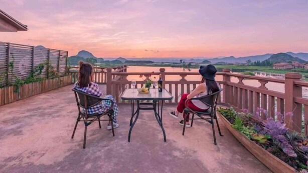 To personer sidder ved et bord på en balkon med udsigt over et smukt landskab med bjerge og vand ved solnedgang.