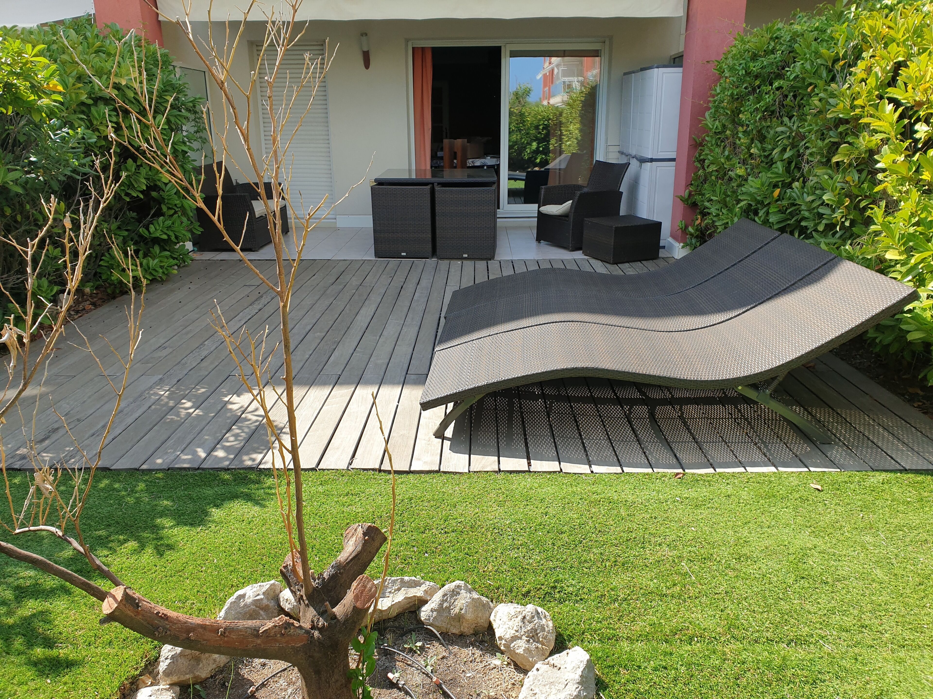 Eine Terrasse mit Esstisch und Stühlen, ein kleiner Gartenbereich mit Steinen und einem Baum sowie ein Gebäude mit einer Schiebetür im Hintergrund.