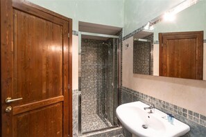 Une salle de bain avec une porte en bois, une douche aux murs carrelés et un lavabo blanc avec un miroir au-dessus.