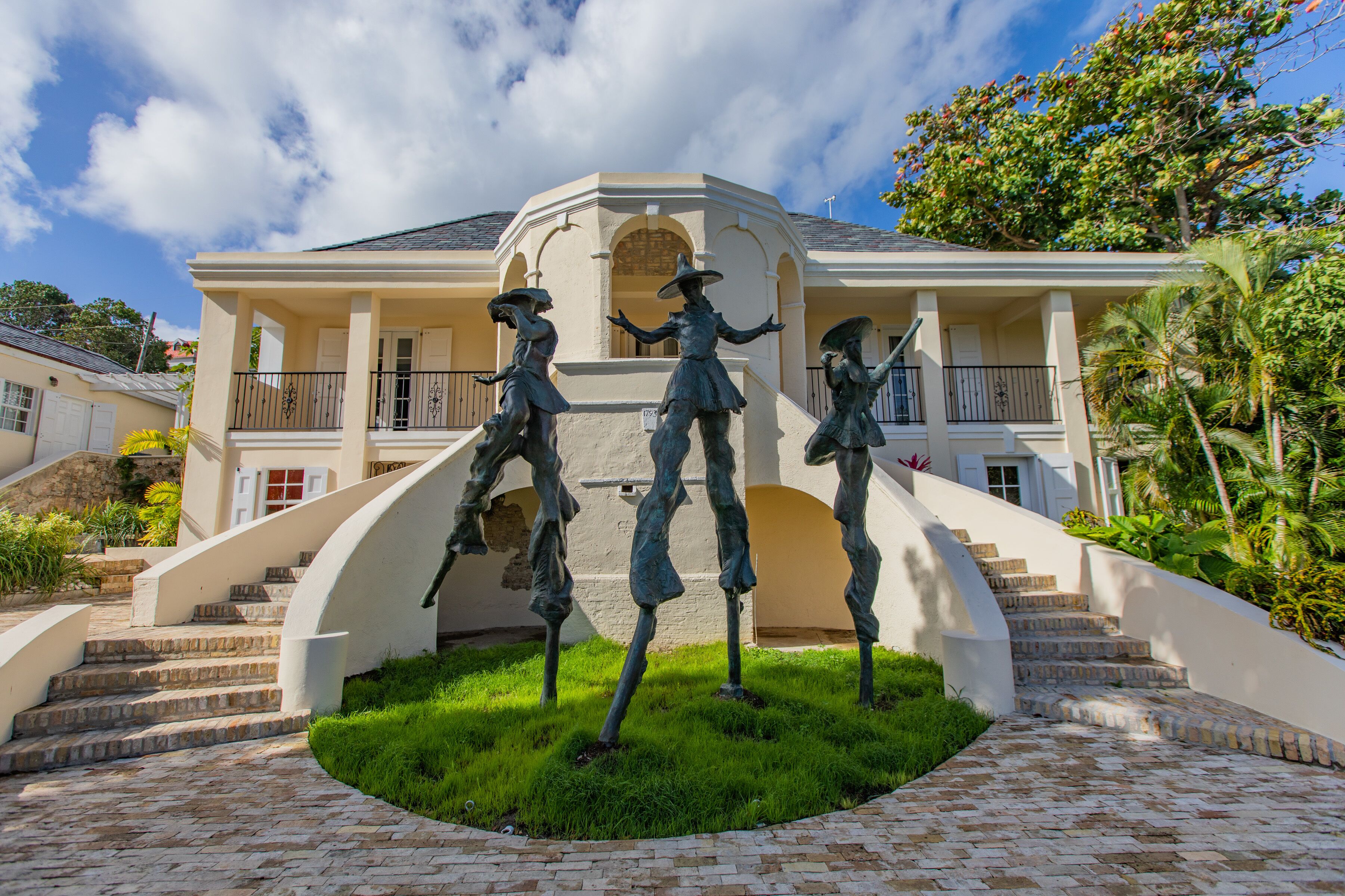 Un edificio con una escalera central, rodeado por una zona circular cubierta de césped y tres estatuas.