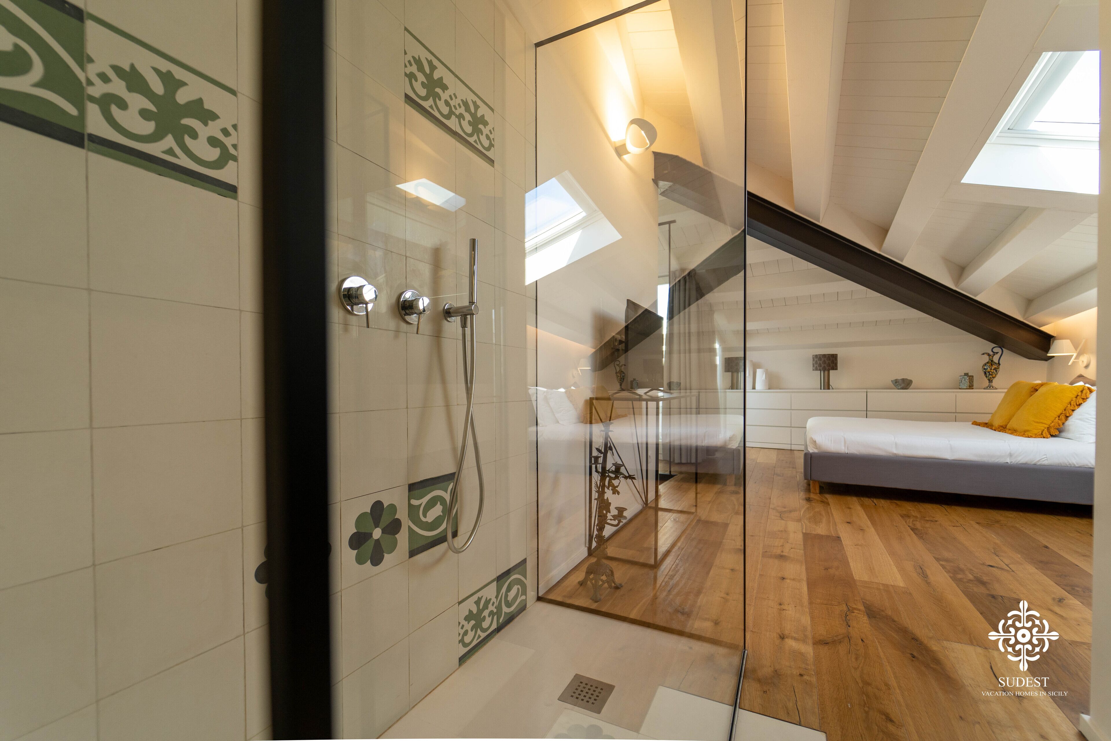 A modern bedroom with a glass shower enclosure, wooden flooring, and a sloped ceiling with skylights.
