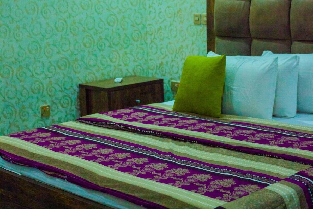 A hotel room with a patterned wallpaper, a bed with a headboard, a nightstand, an air conditioning unit, and a framed picture on the wall.