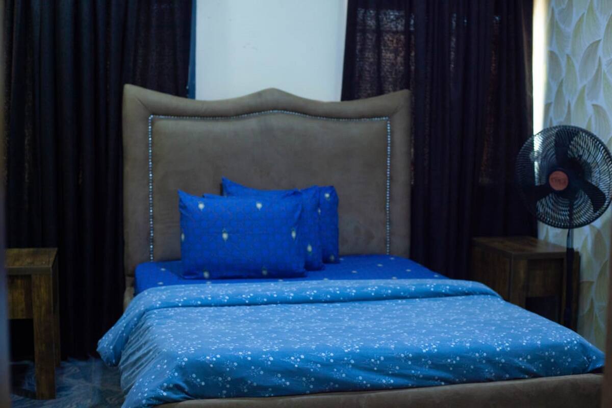 A bedroom with a bed, blue bedding, a headboard, a nightstand, and a standing fan.