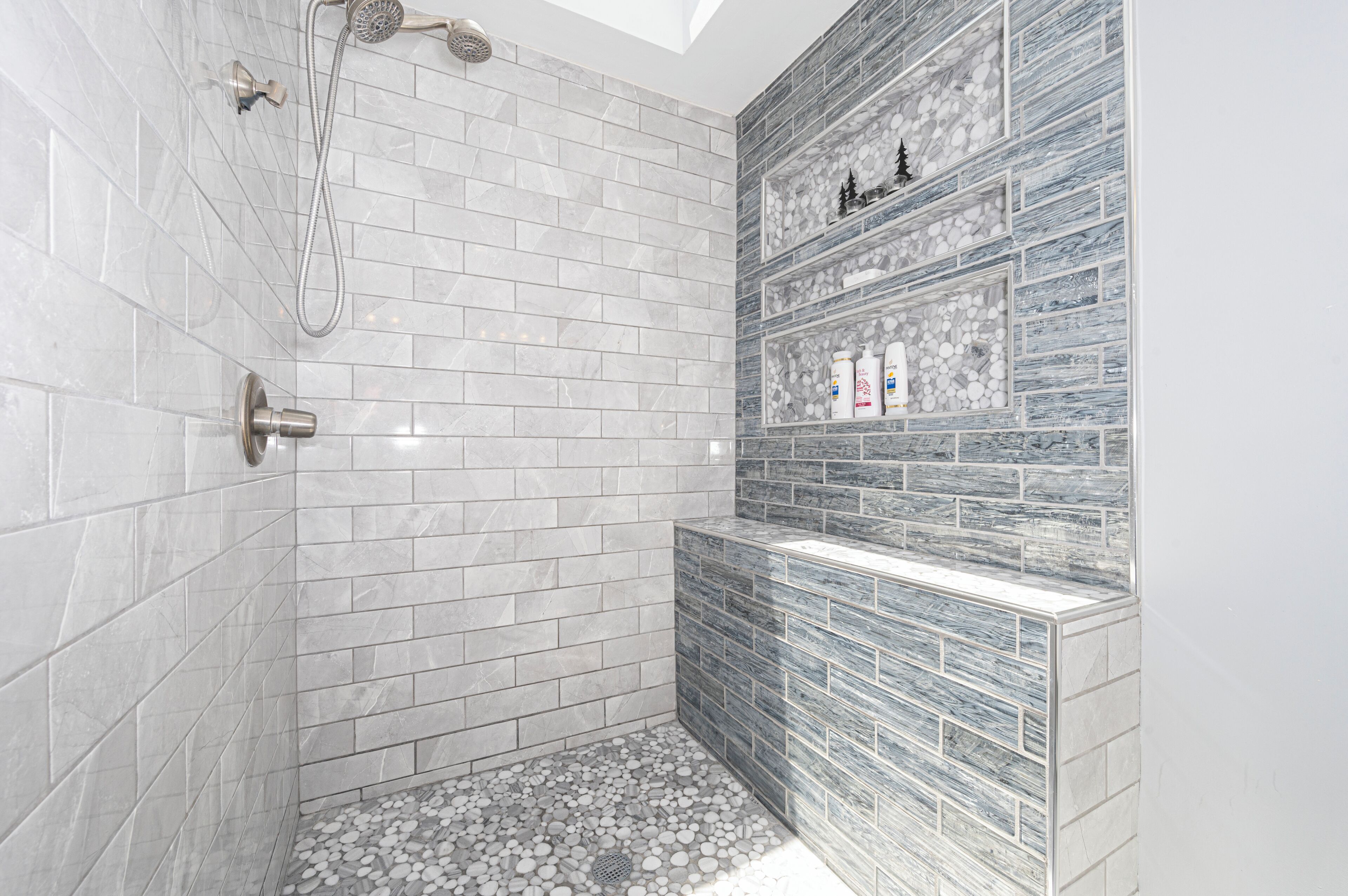 Ein modernes Badezimmer mit weißen und grauen Marmorfliesen, einer Dusche inklusive Handbrause und einem Ablagefach für Toilettenartikel.