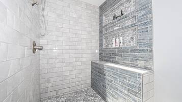 Ein modernes Badezimmer mit weißen und grauen Marmorfliesen, einer Dusche inklusive Handbrause und einem Ablagefach für Toilettenartikel.