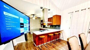 Een moderne keuken met een centraal eiland, barkrukken en een groot scherm waarop een streamingdienst draait.