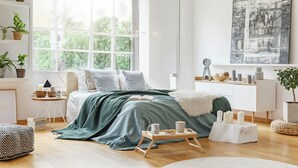 Een moderne slaapkamer met een groot raam, een bed met een groene sprei, nachtkastjes met kaarsen, en een abstract schilderij aan de muur.