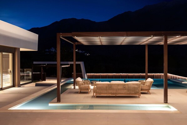 Uma área de piscina moderna ao ar livre, com um espaço de estar coberto e uma sala com paredes de vidro.