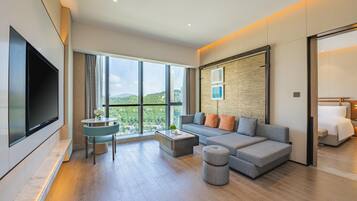 A modern living room with a large flat-screen TV, a grey sofa with orange cushions, a round coffee table, and a view of greenery through large windows.